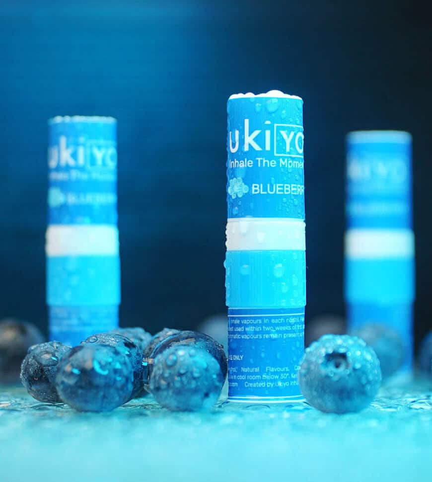 Three "ukiyo" blueberry-flavored inhalers standing upright, with fresh blueberries on a reflective surface against a blue background.