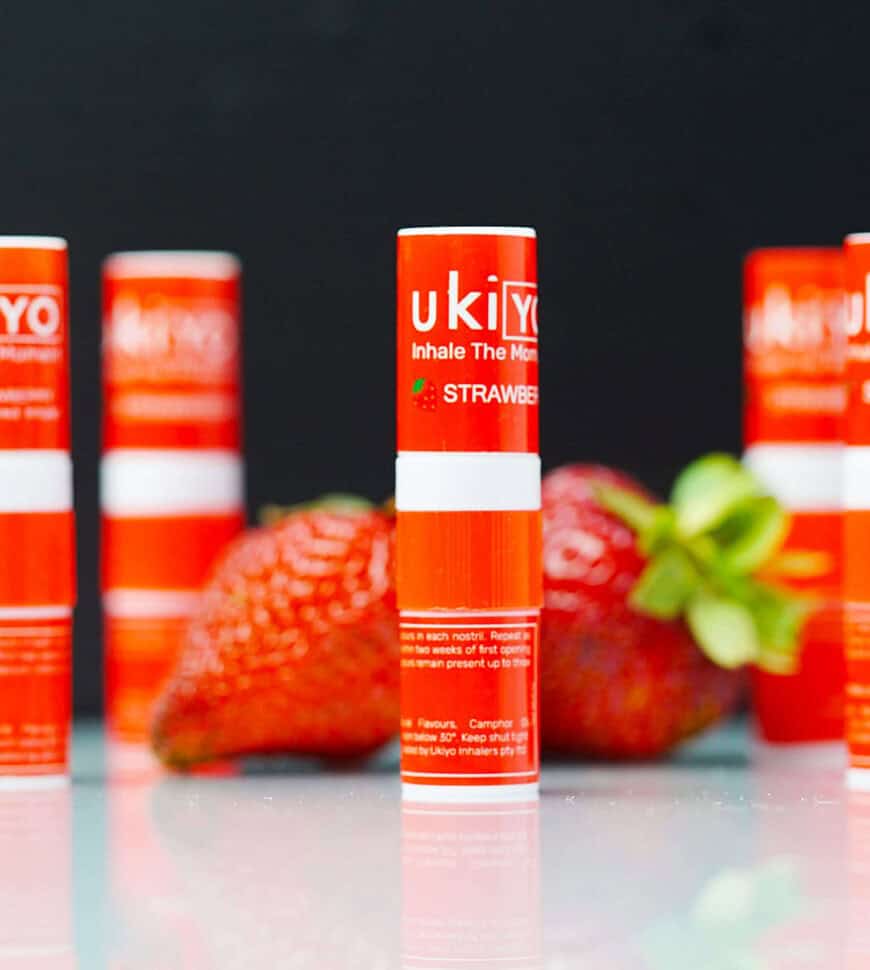 A close-up of a strawberry-flavored ukiyo nasal inhaler with blurred inhalers and strawberries in the background.