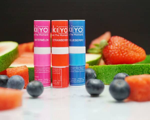 Three lip balms labeled "Mixed Twelve Pack" in watermelon, strawberry, and blueberry flavors, displayed in front of corresponding sliced fruits on a reflective surface.