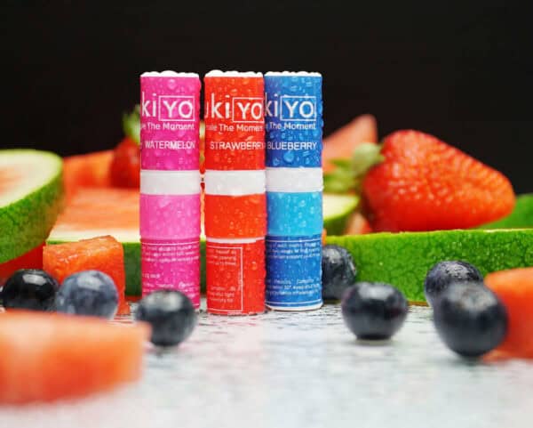 Three colorful lip balms displayed in front of a Mixed Twelve Pack and scattered strawberries and blueberries.