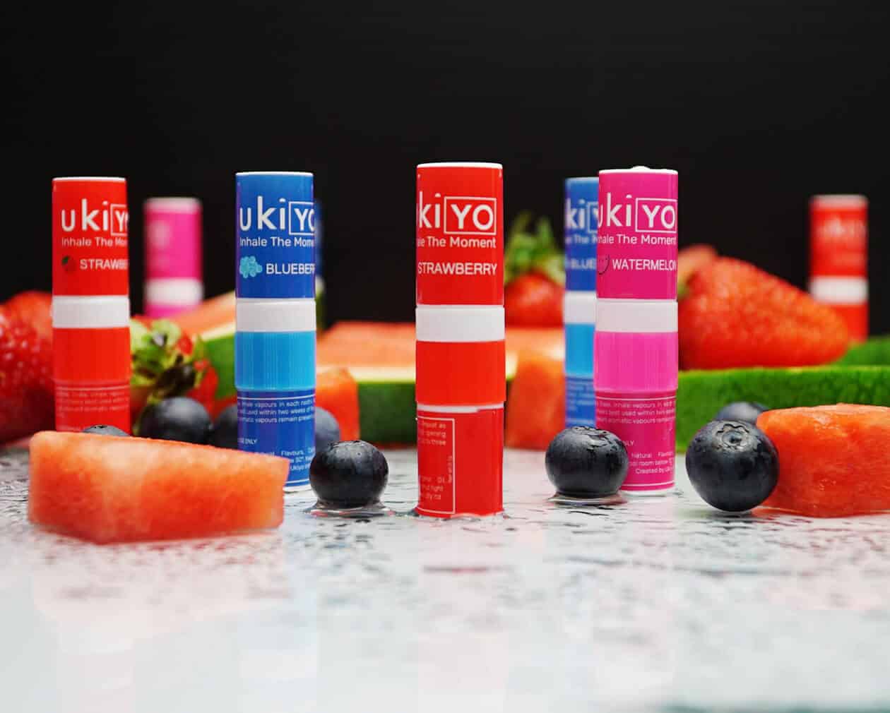 Several Mixed Twelve Pack inhaler tubes arranged with fresh strawberries, blueberries, and watermelon slices on a dark surface.