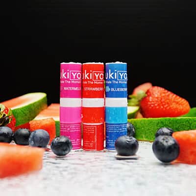 Three lip balms with fruit designs, labeled kiyo kiyo, flanked by fresh fruits including watermelon, strawberries, and blueberries on a dark surface.