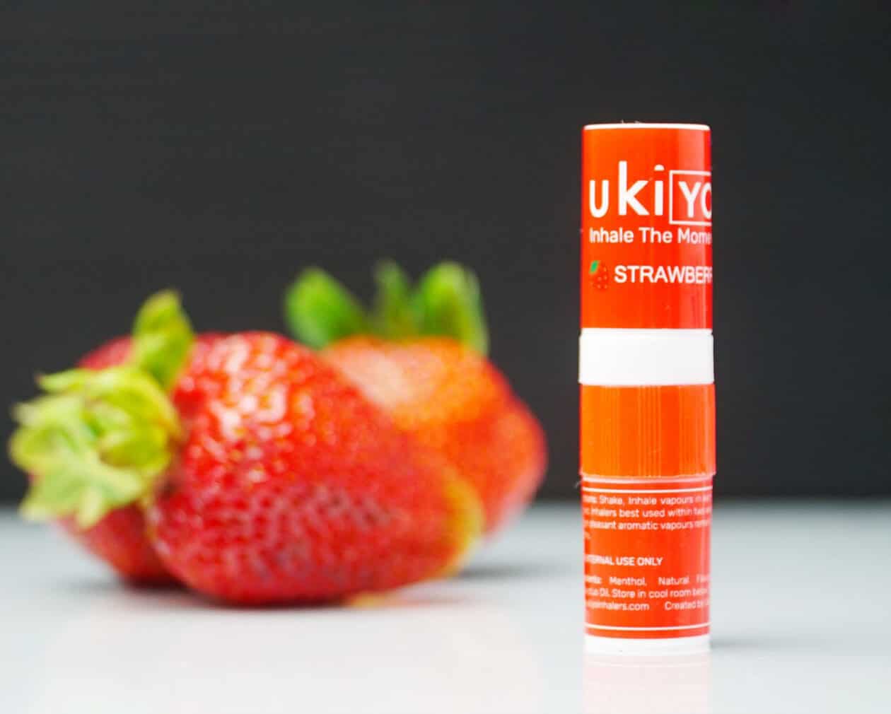 A red lip balm tube labeled "Strawberry Twelve Pack" stands on a white surface with strawberries in the background.