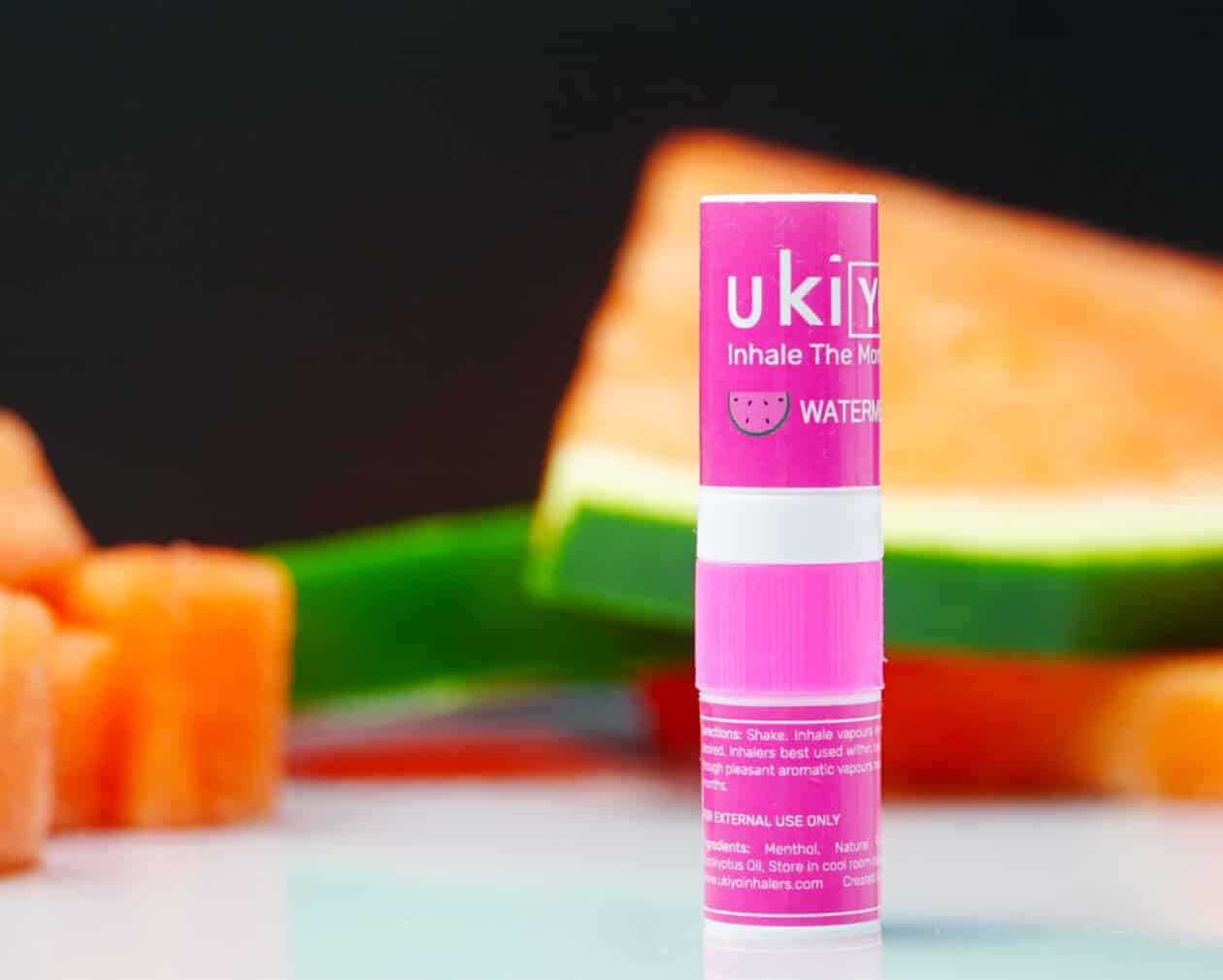 Close-up of a pink lip balm labeled "Watermelon Single Pack" against a backdrop featuring watermelon slices and orange cubes.