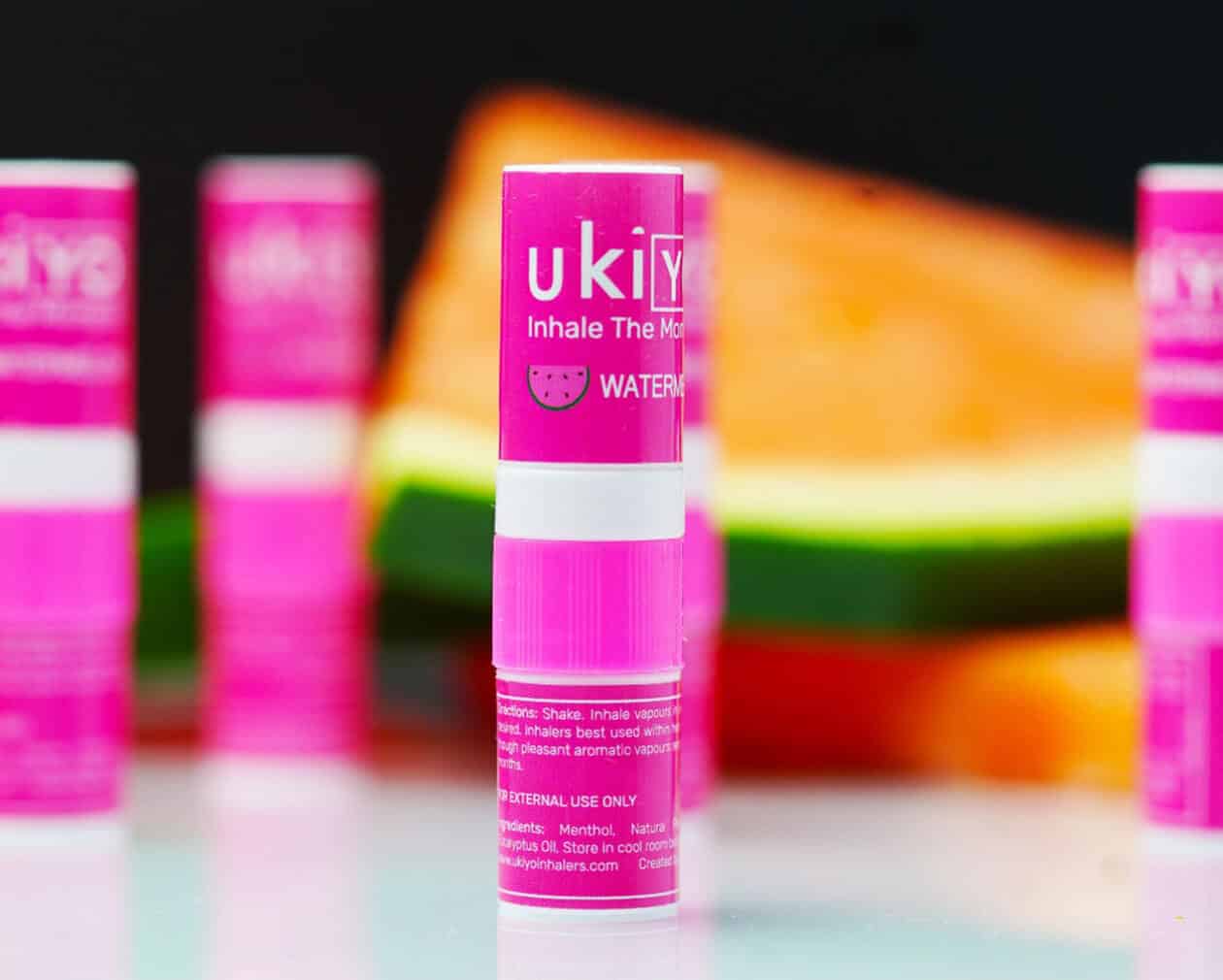 Several pink lip balm tubes labeled "Watermelon Triple Pack" are on a sleek surface with watermelon slices in the background.