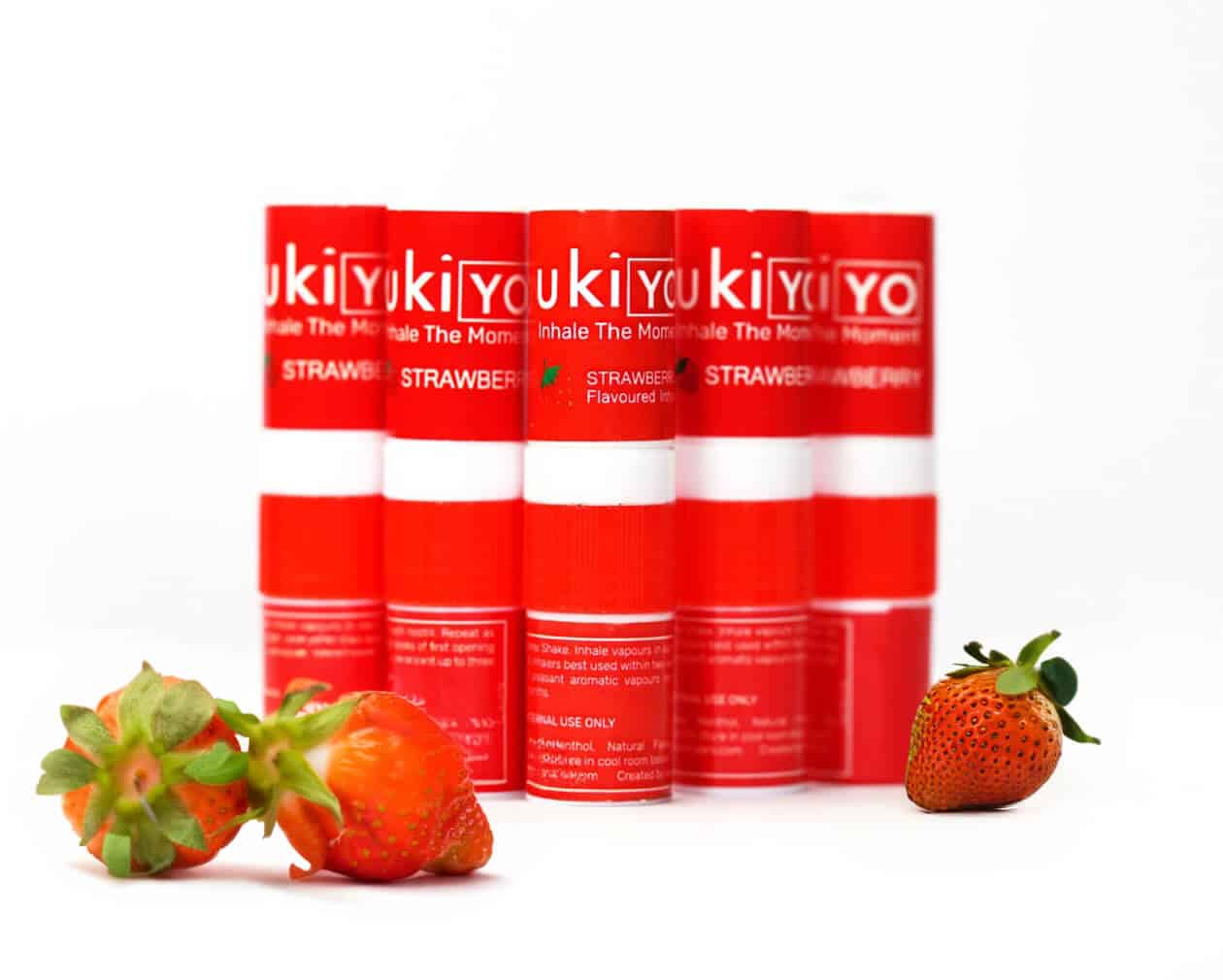 Five red and white cylindrical strawberry-flavored inhalers are lined up, with three strawberries laying in front of them.